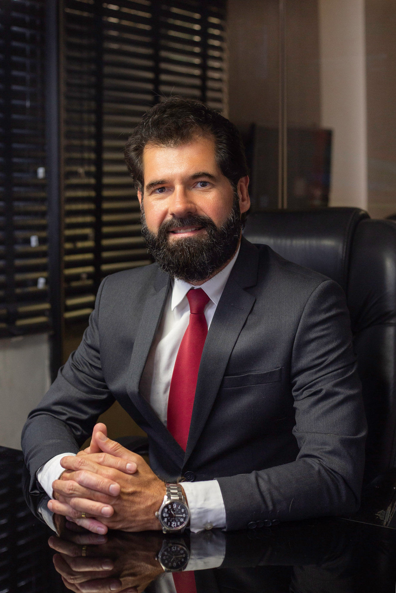 Adriano Sobreira sorri para a câmera. Ele é um homem branco, de cabelo, barba e bigode curtos e pretos, e usa um terno preto. Está sentado em uma cadeira de escritório.