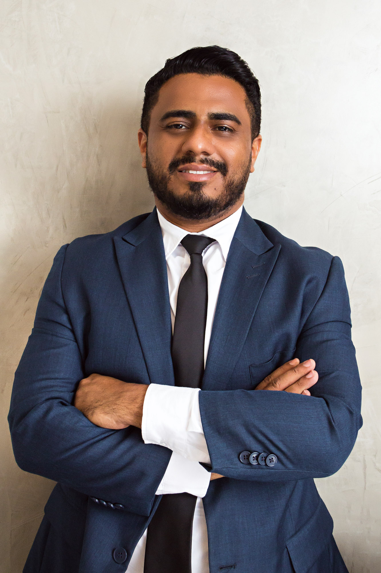 George é um homem negro, jovem, que sorri para a câmera de braços cruzados. Ele tem cabelo, barba e bigode curtos e escuros e usa um terno azul escuro com camisa social branca.