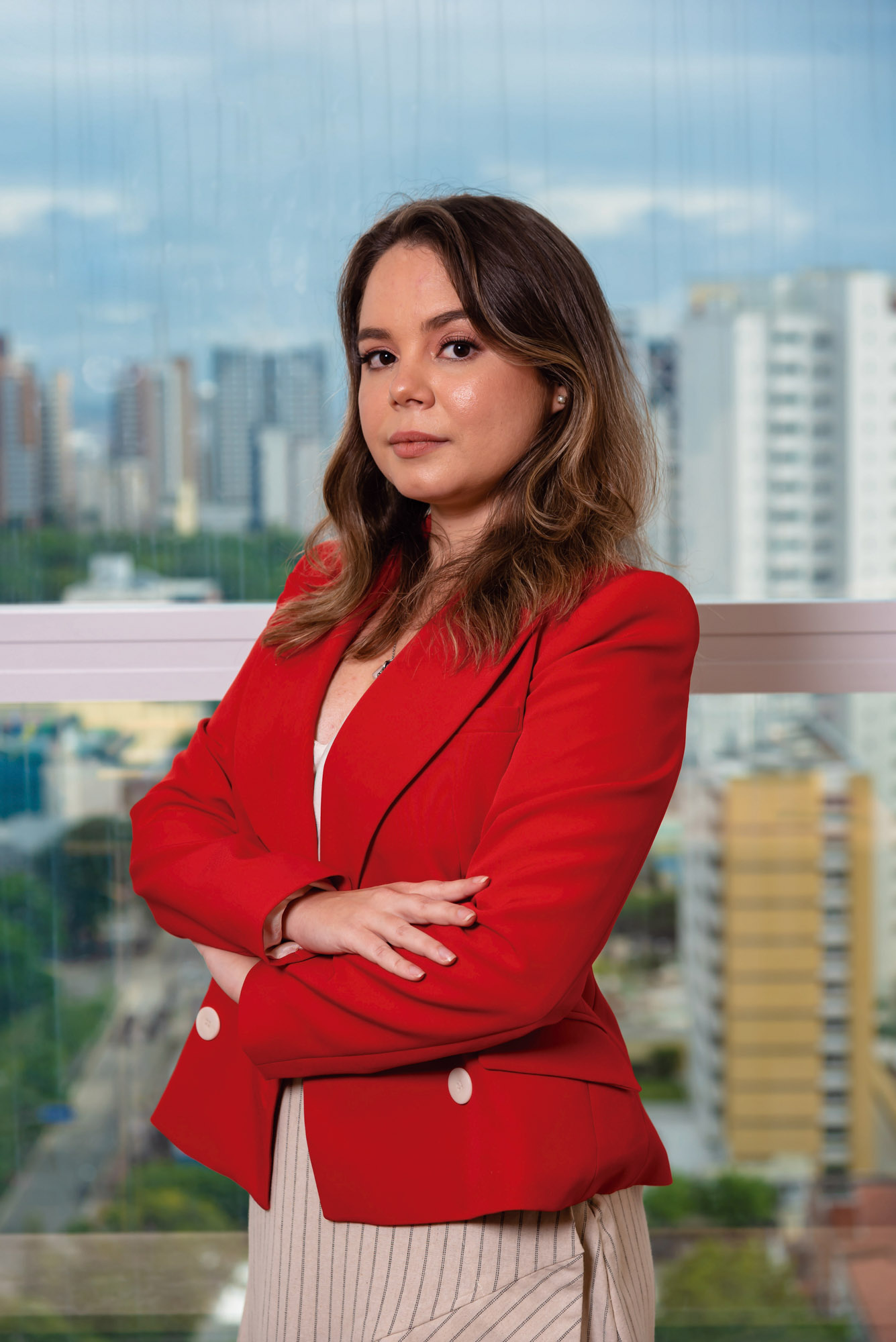 Jéssica é uma mulher branca e jovem, de cabelos castanhos abaixo do ombro. Ela está de braços cruzados e usa blazer vermelho.