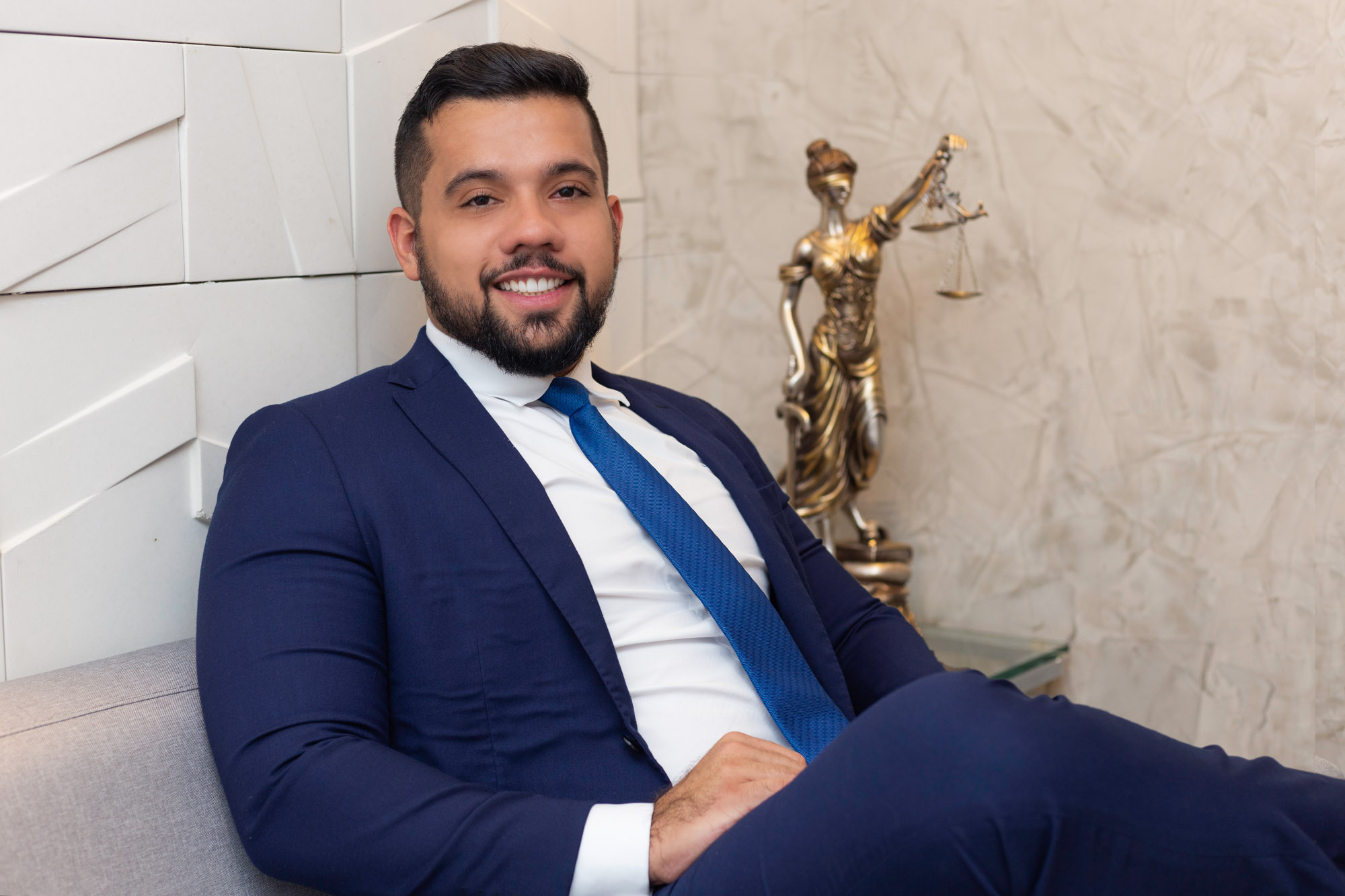 José Aurélio é um homem branco, jovem, de cabelo, barba e bigode curtos e escuros. Ele está sentado em frente a estátua que representa a Justiça e usa terno e gravata azuis.