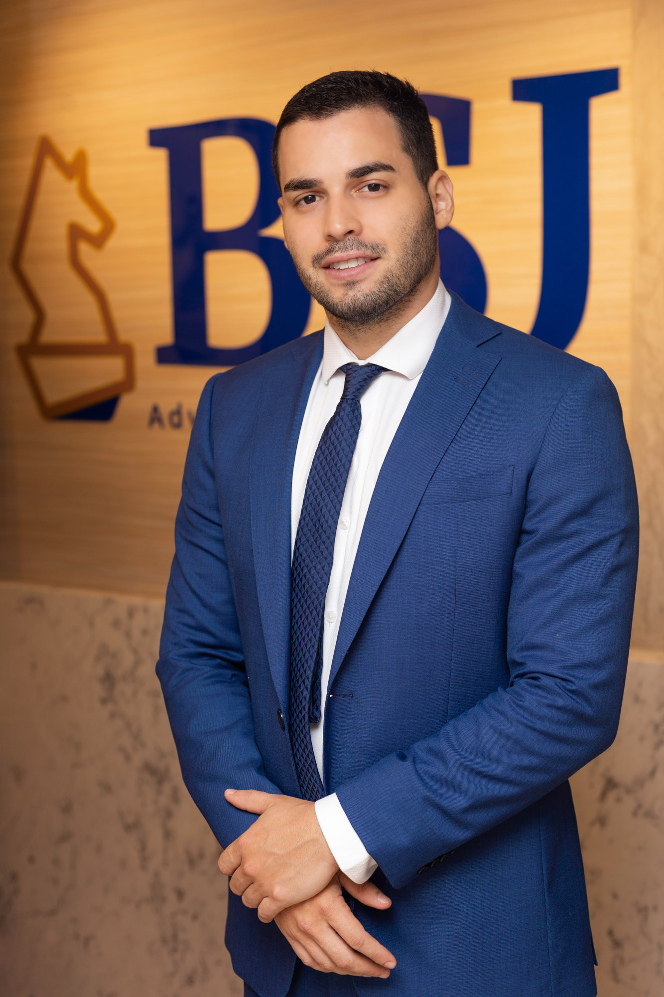 Victor sorri em frente ao logo do escritório BSJ Advogados. Ele é um homem branco, jovem, de cabelo, barba e bigode escuros e curtos. Ele usa um terno azul.