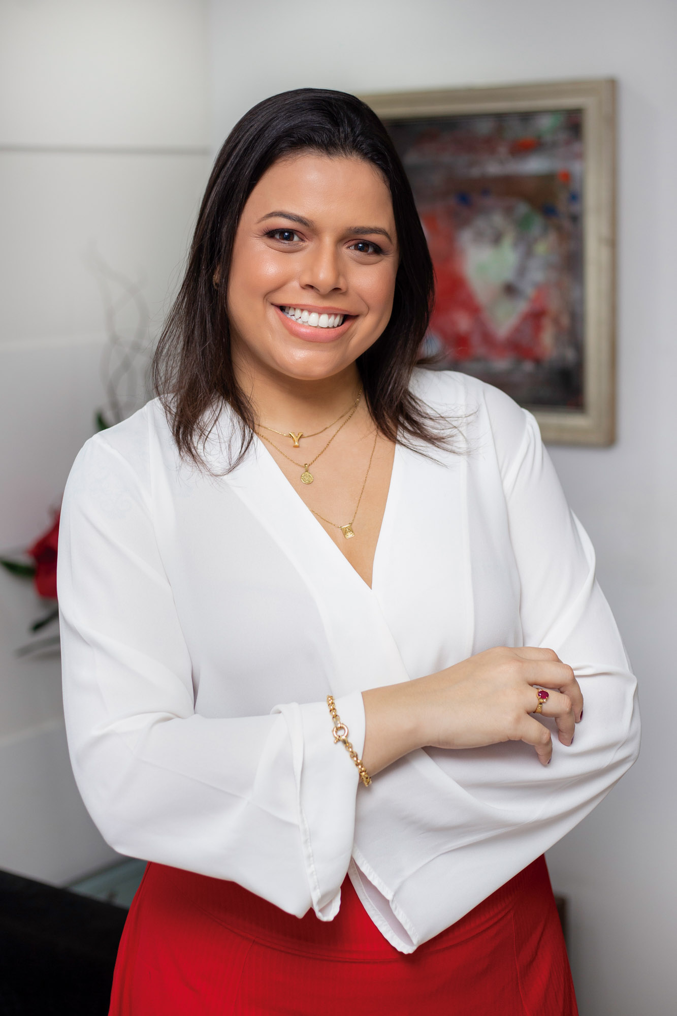 Yáskara sorri para a câmera em frente a um quadro. Ela é uma mulher jovem, de cabelos curtos e castanhos, e usa uma camisa social branca.